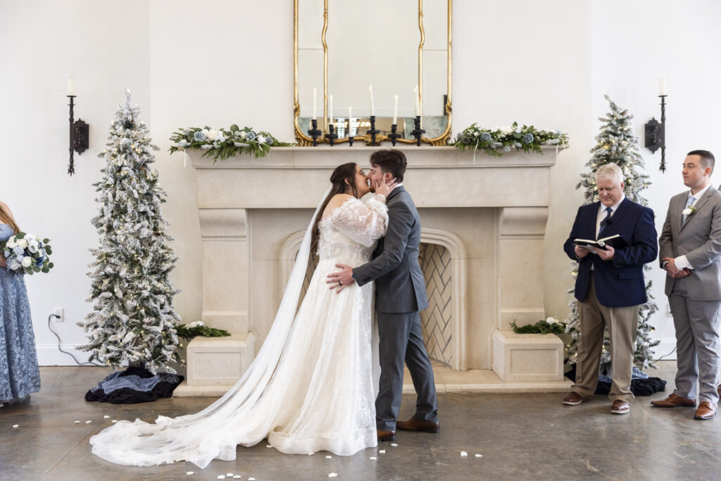 The couple shares their first kiss beneath the mantle, framed by tall mirrors, candles, and winter décor, as guests and the officiant look on in these howe farms wedding photos filled with soft light and classic symmetry.