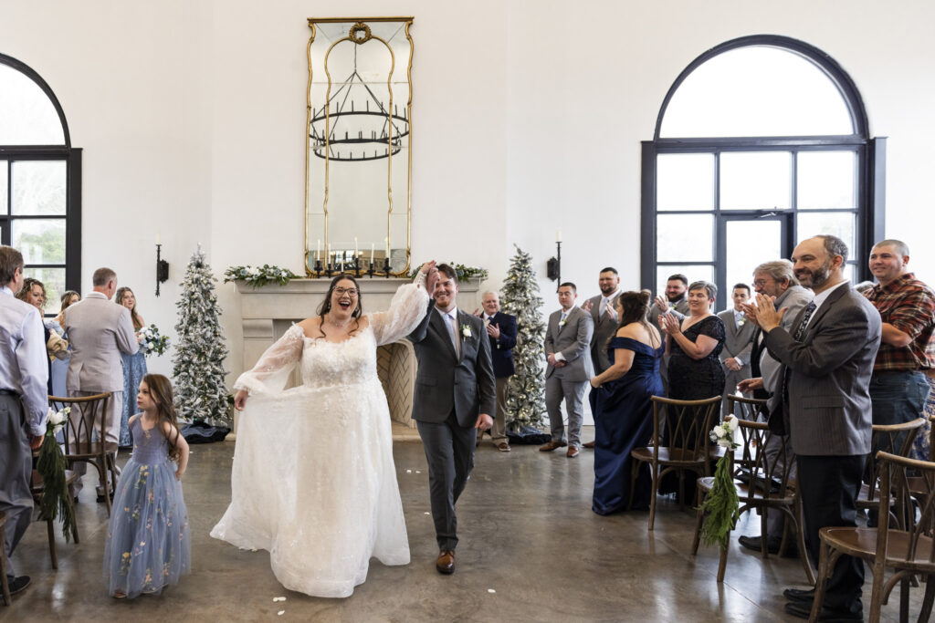 The bride and groom make their joyful recessional down the aisle after the ceremony, hands raised and smiles wide, as guests stand and applaud from wooden chairs on either side of the aisle.