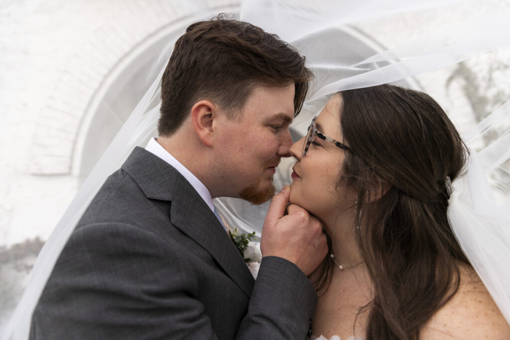 A close-up portrait shows the groom gently lifting the bride’s chin as they lean in nose to nose, smiling softly beneath her flowing veil. The delicate lace of her dress and the groom’s boutonniere are sharply in focus, with the pale brick backdrop of the howe farms wedding venue softly blurred behind them.