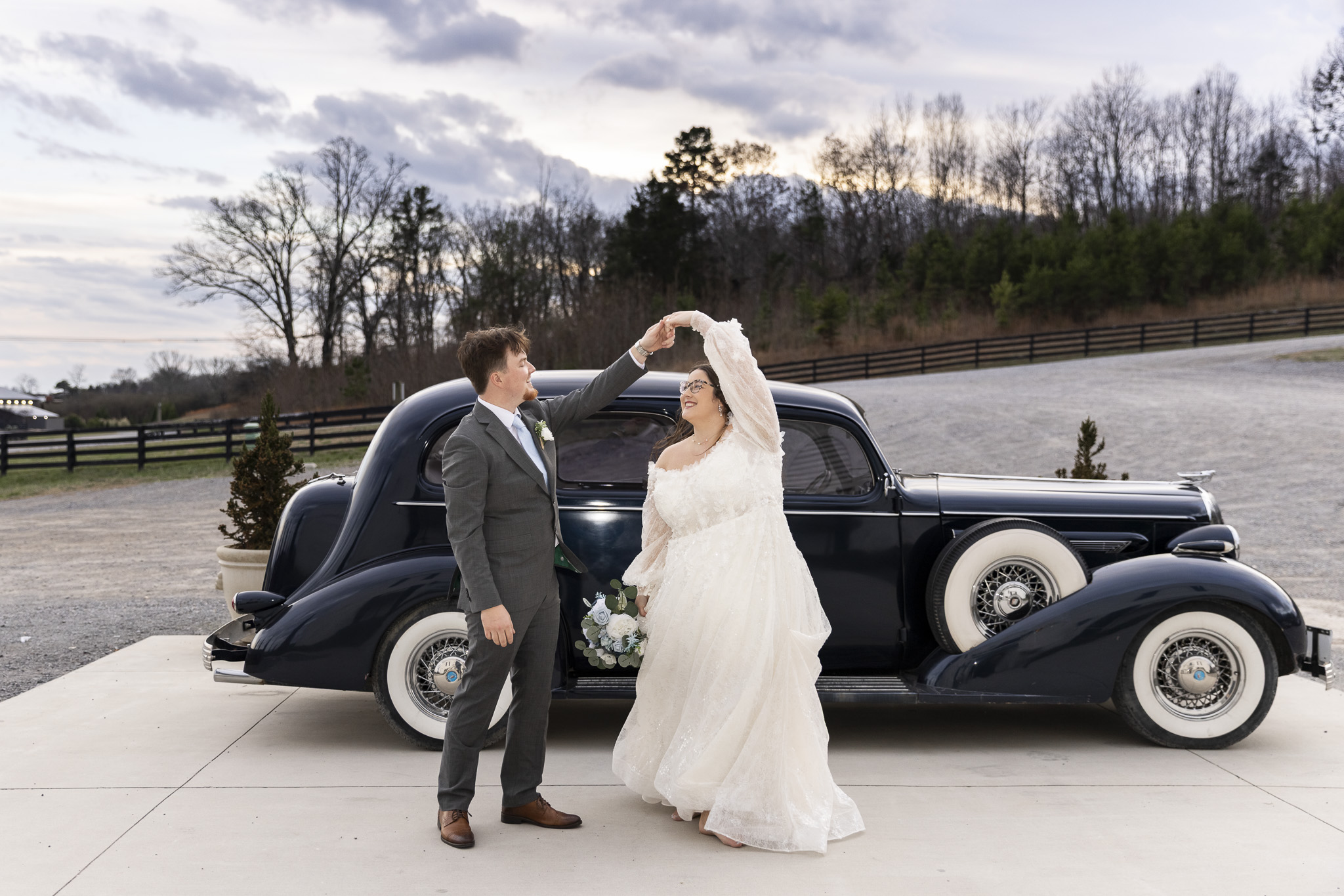 Outside at The Woodlands at Howe Farms, the newlyweds share a playful twirl in front of a vintage black car, the groom lifting the bride’s hand as her full lace skirt moves in the breeze against a backdrop of rolling hills and winter trees.