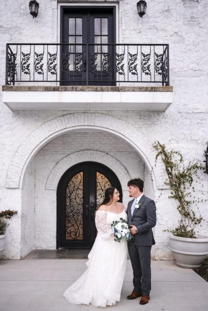 The couple stands close together beneath an arched brick entryway at The Woodlands at Howe Farms, gazing at one another while holding a bouquet of blue and white florals, framed by textured white walls, black doors, and potted greenery.