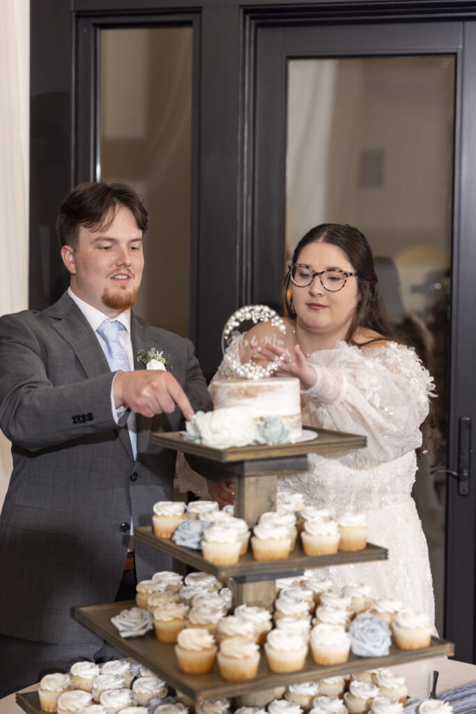 The couple stands side by side at their dessert display, gently cutting into a small round cake placed atop a tiered stand of frosted cupcakes, with glass doors and softly glowing interior lights creating a cozy reception backdrop.