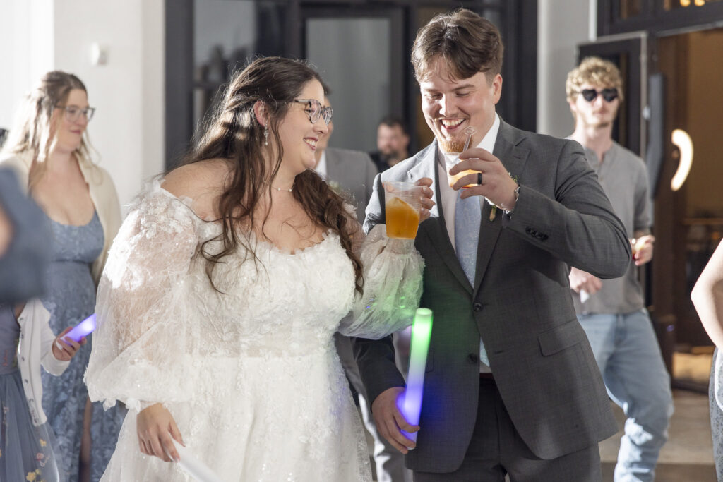 On the dance floor, the bride and groom laugh while holding plastic cups filled with amber-colored drinks, surrounded by guests waving glowing light sticks. The bride’s lace sleeves catch the colorful light as the celebration unfolds inside the howe farms wedding venue during the reception.