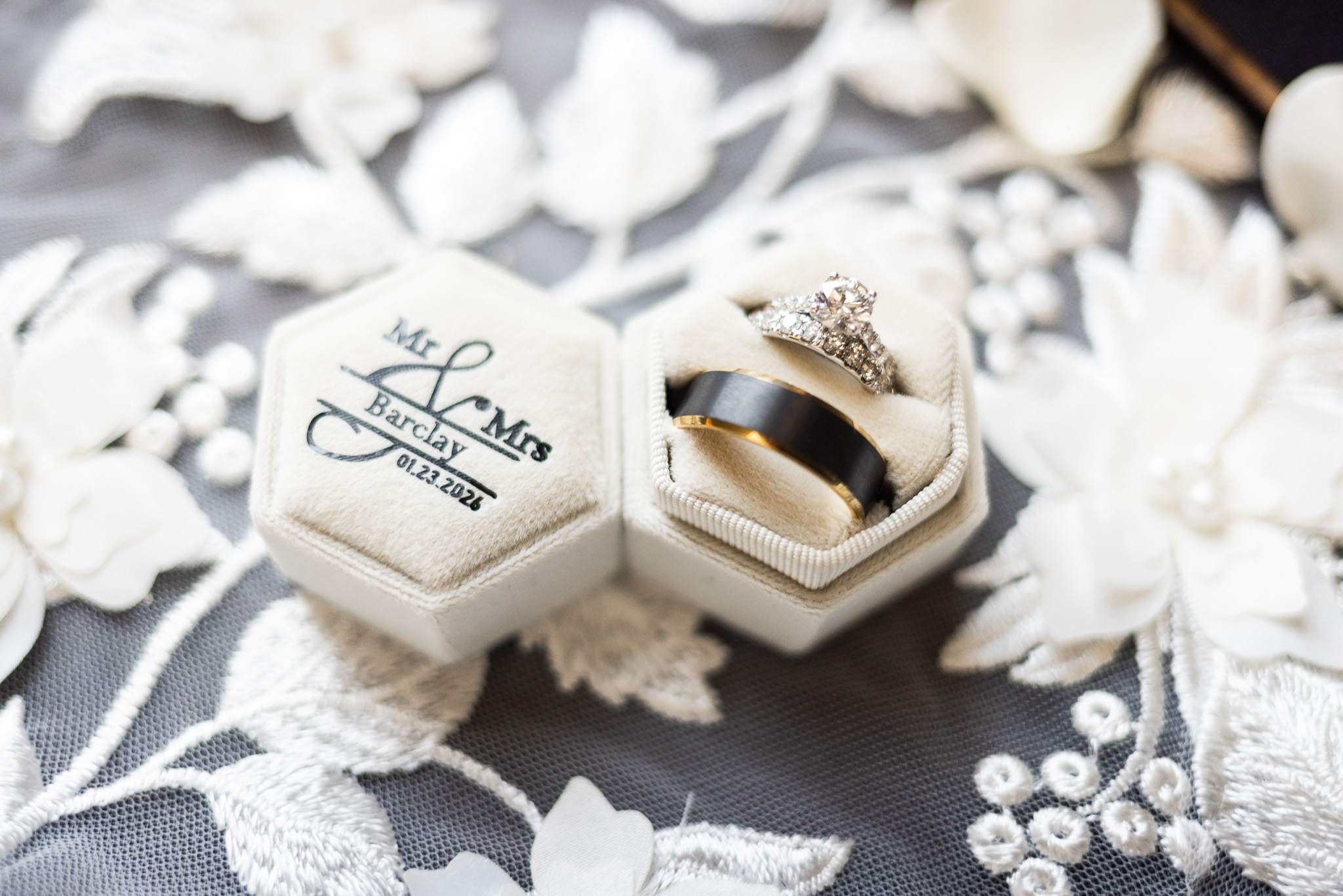 Close-up flat lay of an open hexagon ring box embroidered with the couple’s last name and wedding date, holding a diamond engagement ring and wedding bands, placed on embroidered lace fabric with pearl accents and soft floral textures surrounding the box.