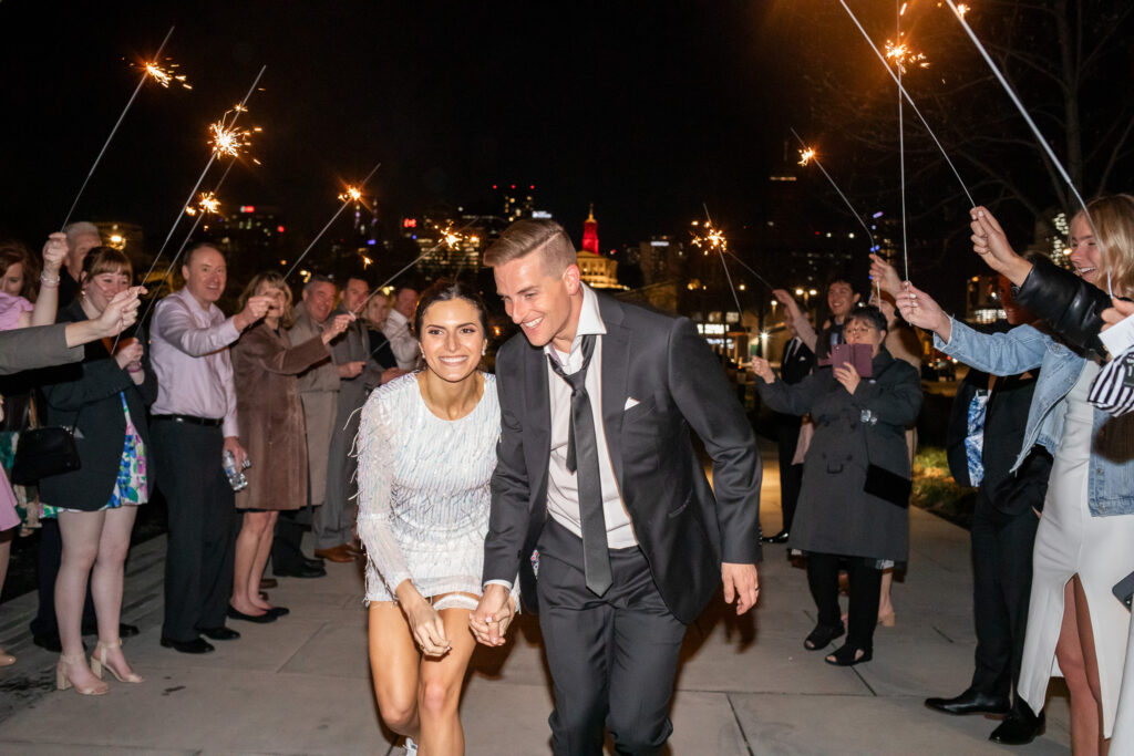 A bride in a short, shimmering reception dress and white sneakers runs hand-in-hand with her groom through a tunnel of guests holding lit sparklers at night, city lights glowing behind them as friends cheer and raise their arms on either side.