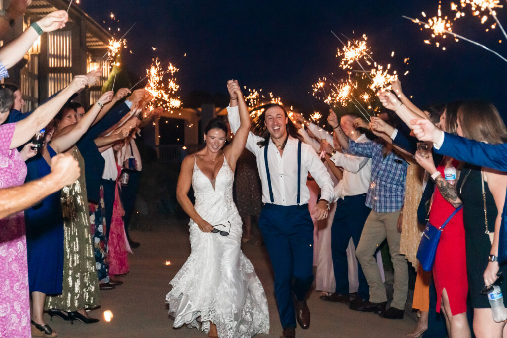 A bride in a fitted lace gown and a groom in suspenders sprint forward with their arms raised triumphantly through a sparkler-lined walkway at night, guests on both sides smiling, holding drinks, and lifting sparklers high to create a glowing arch overhead.