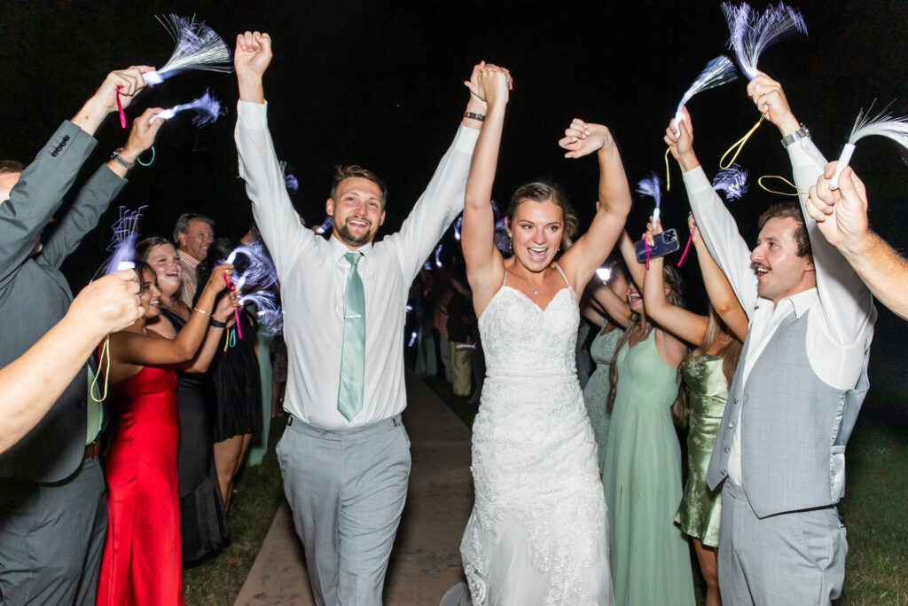 A bride in a lace wedding gown and a groom in a light gray vest and tie walk forward with their hands raised, surrounded by guests waving glowing fiber-optic wands that create soft streaks of white light against the dark night sky.