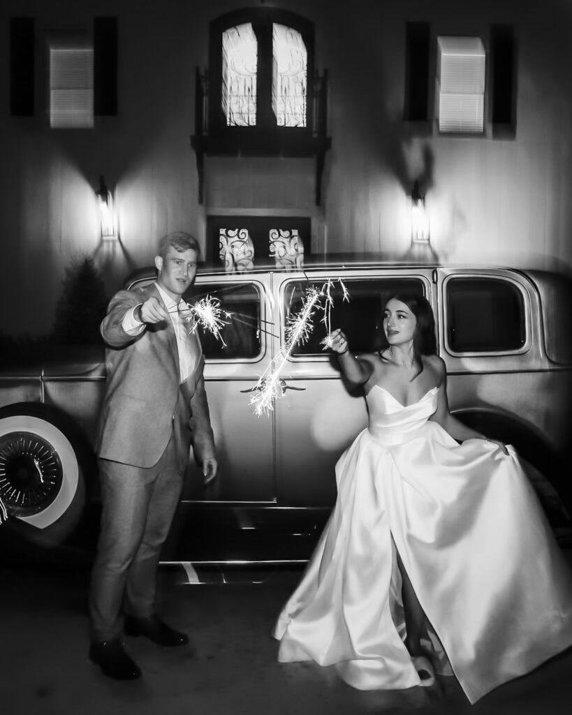 In a dramatic black-and-white portrait, a bride in a structured satin gown and a groom in a tailored suit hold sparklers beside a vintage car parked in front of a building with tall windows, the bright sparks illuminating their faces against the dark evening backdrop.