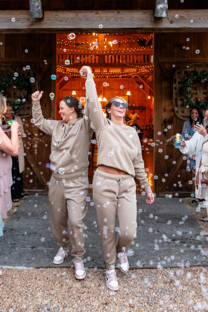 Two brides in matching tan sweatsuits and sneakers run out of a rustic barn doorway laughing with their hands raised, bubbles floating around them while friends cheer on both sides, creating a playful and relaxed wedding exit moment.