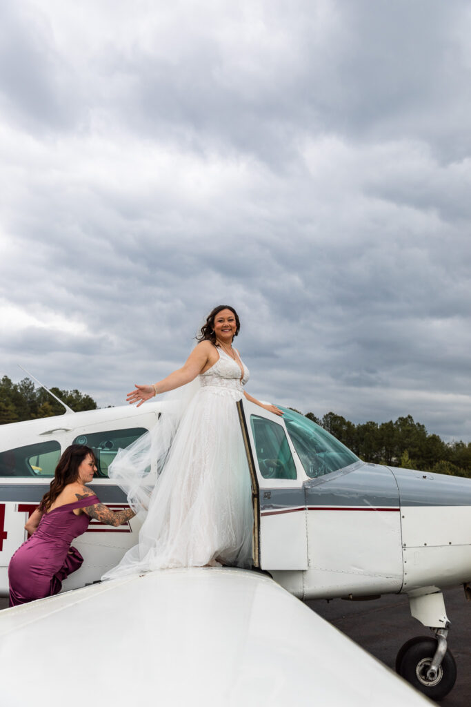 Under a cloudy daytime sky, a bride stands smiling on the wing of a small white airplane with her arms open wide while another woman steadies her gown, capturing a bold and adventurous take on a wedding exit before takeoff.