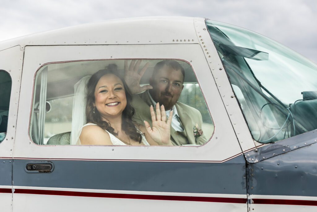 A bride and groom sit inside a small airplane cockpit, smiling and waving through the window as if departing for their honeymoon, the bride’s veil visible and the groom seated behind her, both framed by the curved aircraft window.