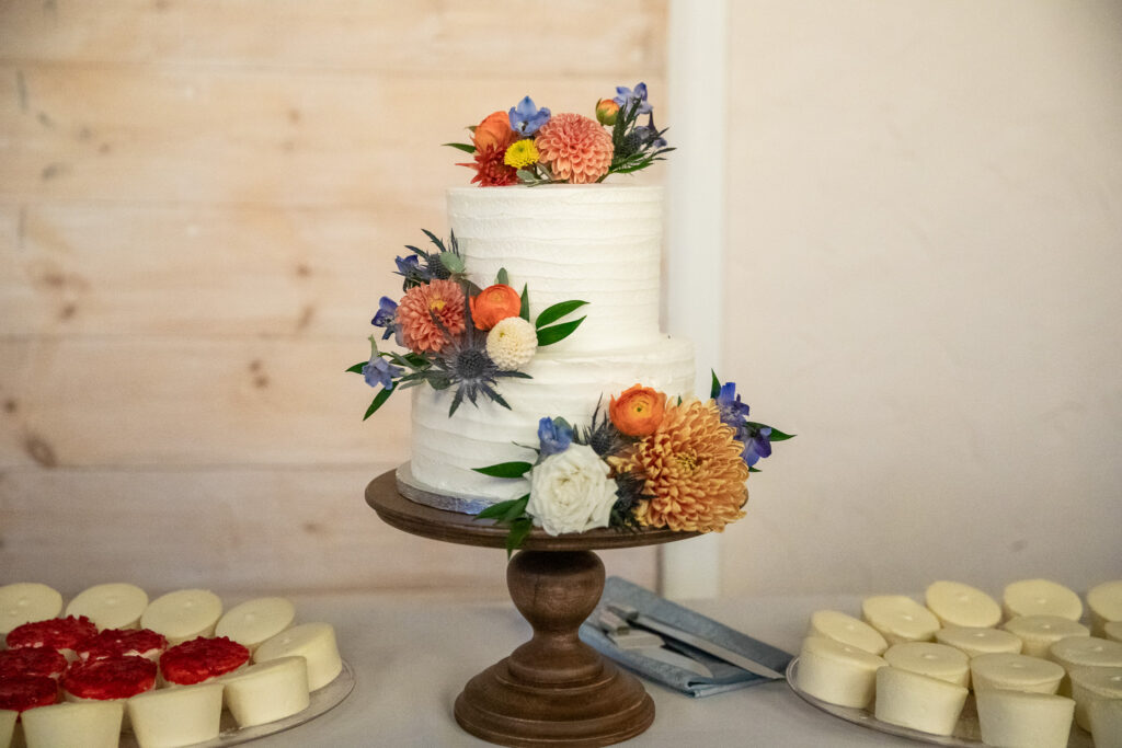 A three-tier white wedding cake displayed on a wooden cake stand, decorated with textured buttercream and clusters of fresh florals in soft orange, blue, and cream tones. Small dessert cups are arranged neatly on either side of the cake on the reception table.
