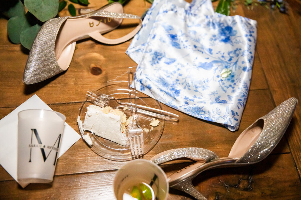 A late night flat lay on a wooden floor showing the bride’s glittering heels, a slice of wedding cake on a clear plate with a fork, and a blue-and-white patterned fabric face mask. Personalized cups with initials sit nearby, hinting at the celebration winding down.