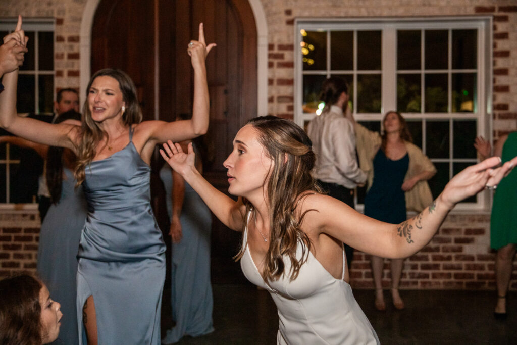 The bride dances enthusiastically on the reception floor with her arms outstretched, surrounded by bridesmaids in soft blue dresses. The moment feels energetic and unfiltered, with guests moving and cheering in the background.