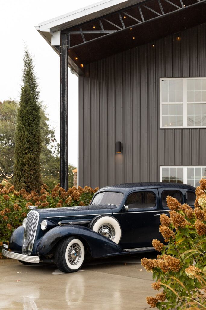 A vintage black car with whitewall tires is parked beside the reception building, surrounded by blooming hydrangeas. The dark exterior of the building and soft landscaping give the scene a timeless, romantic feel.