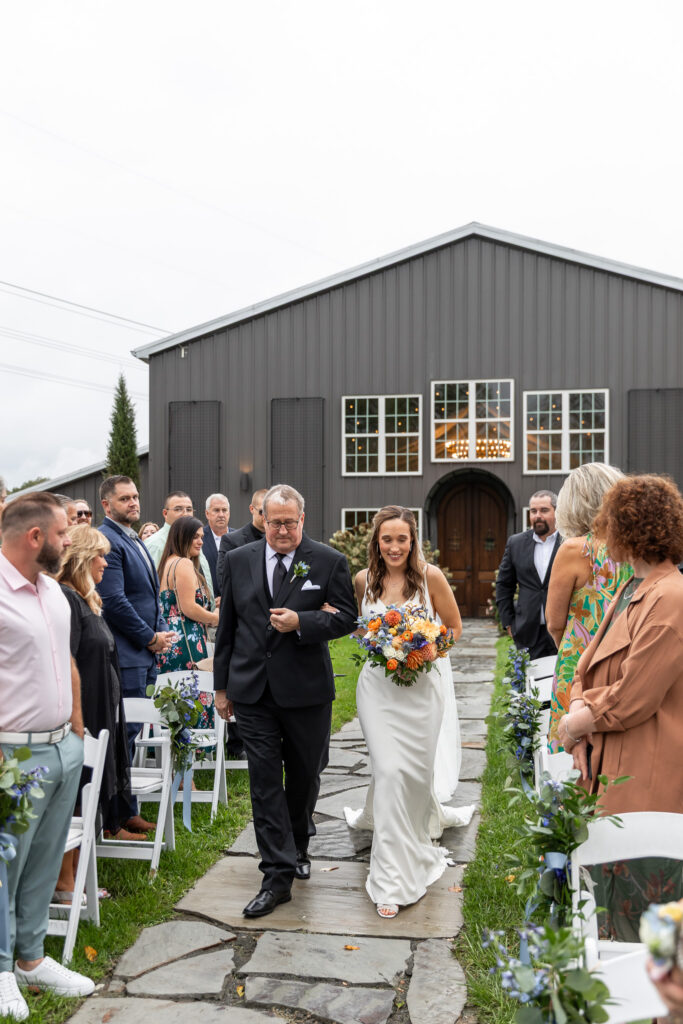 The bride walks down the stone aisle at The Loft at Howe Farms with a loved one at her side, holding a colorful bouquet of orange and blue florals as guests stand along white ceremony chairs lining the path.