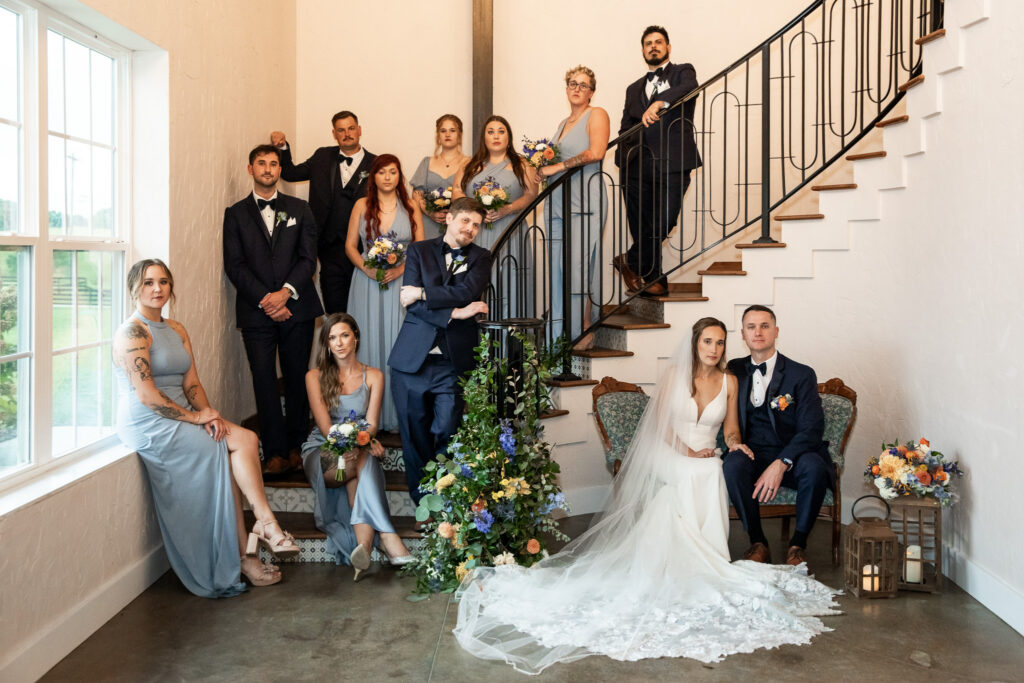 The full wedding party gathers on the staircase inside The Loft at Howe Farms, with the bride and groom seated at the base, their attendants arranged along the steps holding bouquets amid iron railings and floral décor.