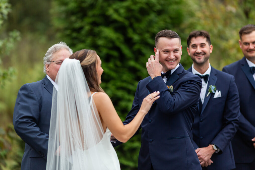 The bride reaches for the groom’s hand during their ceremony at a Chattanooga wedding venue, her veil draped down her back as he smiles and lifts his hand to his face, clearly emotional, with groomsmen standing behind him.