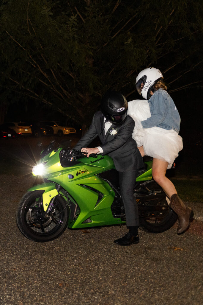 A groom in a tuxedo sits astride a bright green sport motorcycle while the bride climbs on behind him in a short reception dress and denim jacket, her helmet labeled “Bride,” creating an unforgettable motorcycle wedding send off in a dimly lit parking lot.