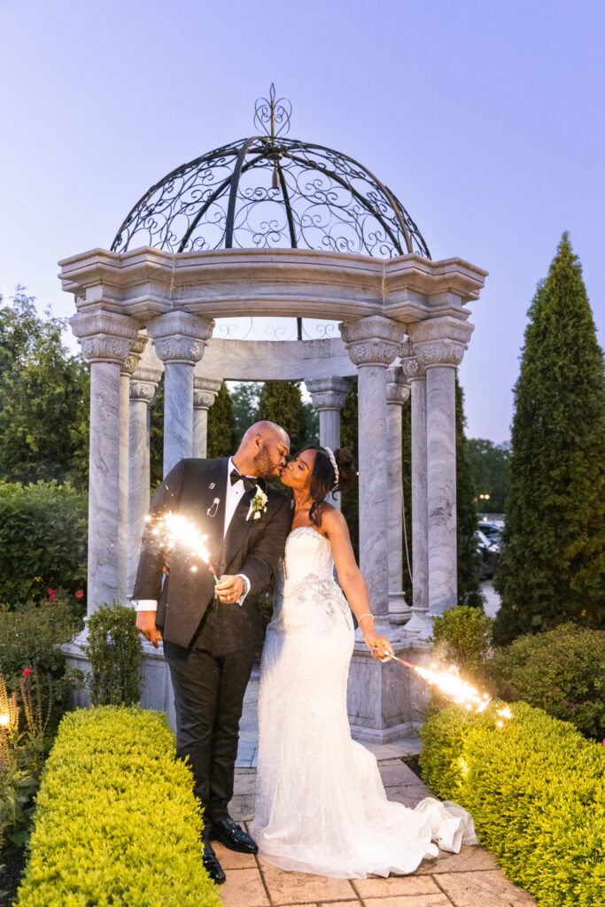 In a manicured garden framed by a stone gazebo with ornate ironwork, the bride and groom lean into a soft kiss while holding lit sparklers at their sides, greenery glowing around them in the early evening light during their romantic wedding exit.