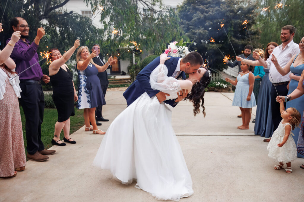 A groom dips his bride into a kiss in the center of a driveway while guests line both sides holding sparklers, a small child in a white dress watching from the foreground as sparks glow warmly against the early evening sky.