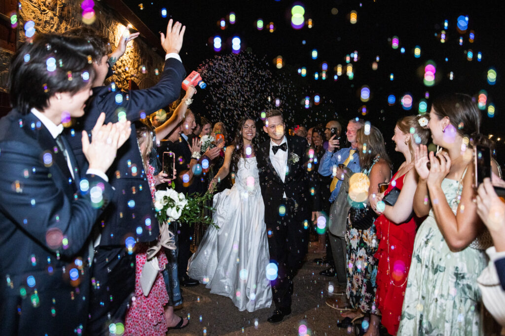 At night outside a stone building, a newly married couple walks arm in arm through a shower of floating bubbles illuminated by flash, colorful light reflections scattered throughout the frame as guests clap and hold bubble guns around them.