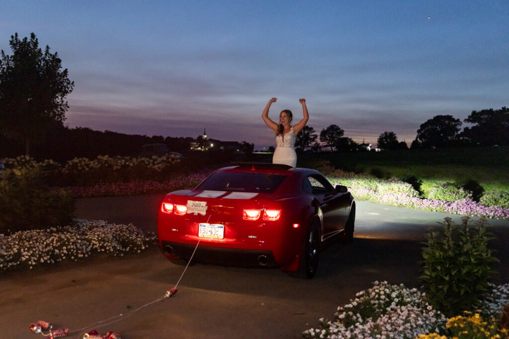 At dusk, a bride stands up through the sunroof of a red sports car decorated with a “Just Married” sign and trailing cans, raising both arms in celebration while the car’s taillights glow red against a darkening countryside landscape.
