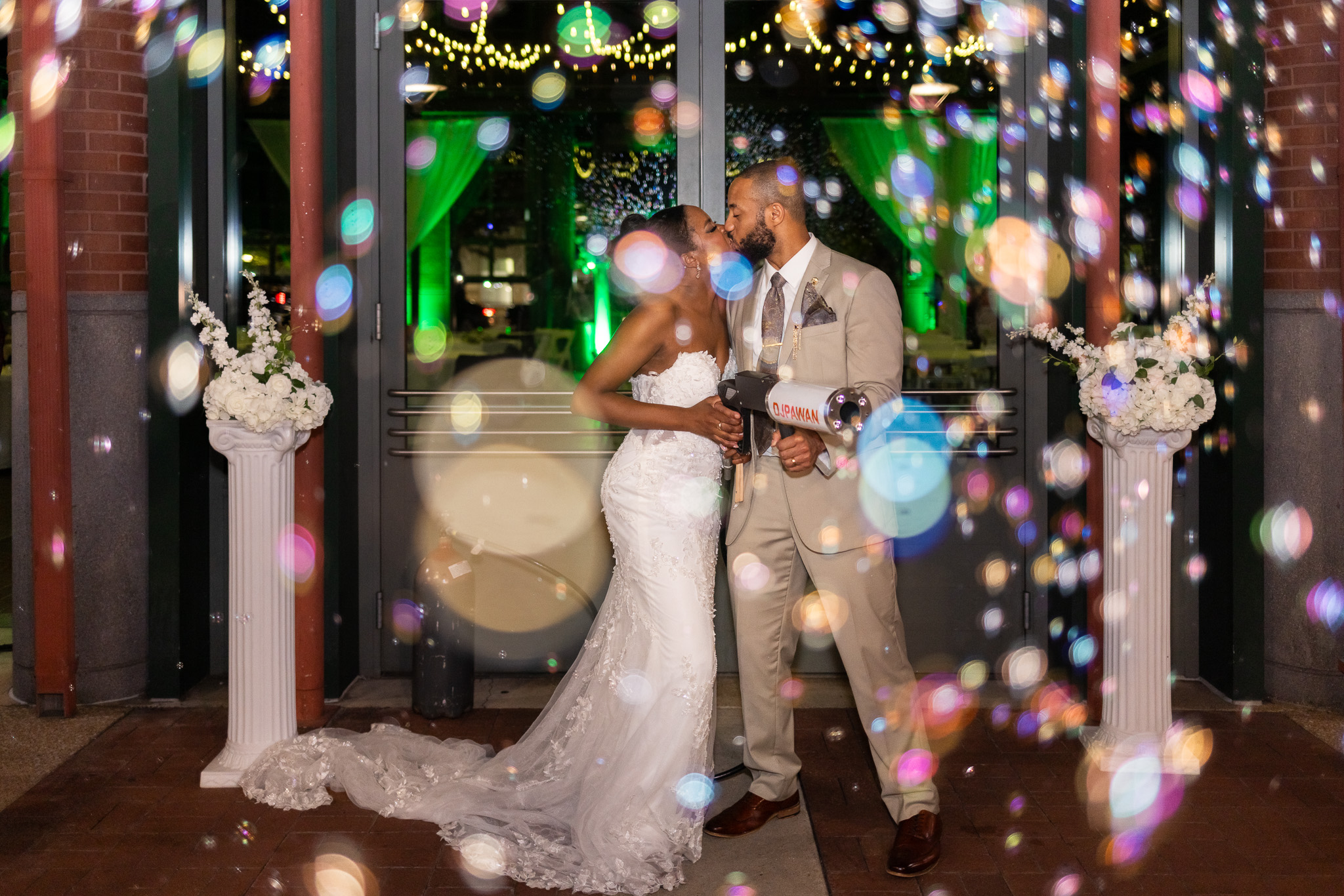 Standing in front of tall glass doors glowing green from uplighting inside, the bride and groom share a kiss while holding a bubble machine between them, colorful reflections dancing across the frame as they pause in the middle of their wedding send off.