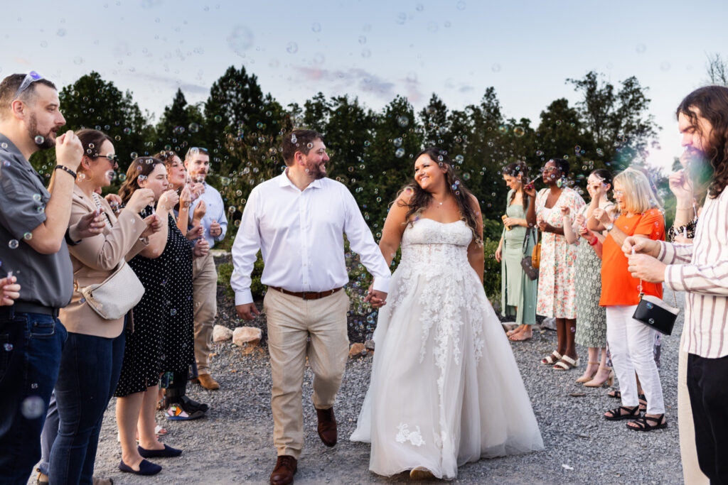 At dusk on a gravel path, guests line both sides blowing bubbles while the bride in a flowing lace gown and the groom in a white shirt walk hand in hand through the center of their wedding send off, smiling softly at one another.