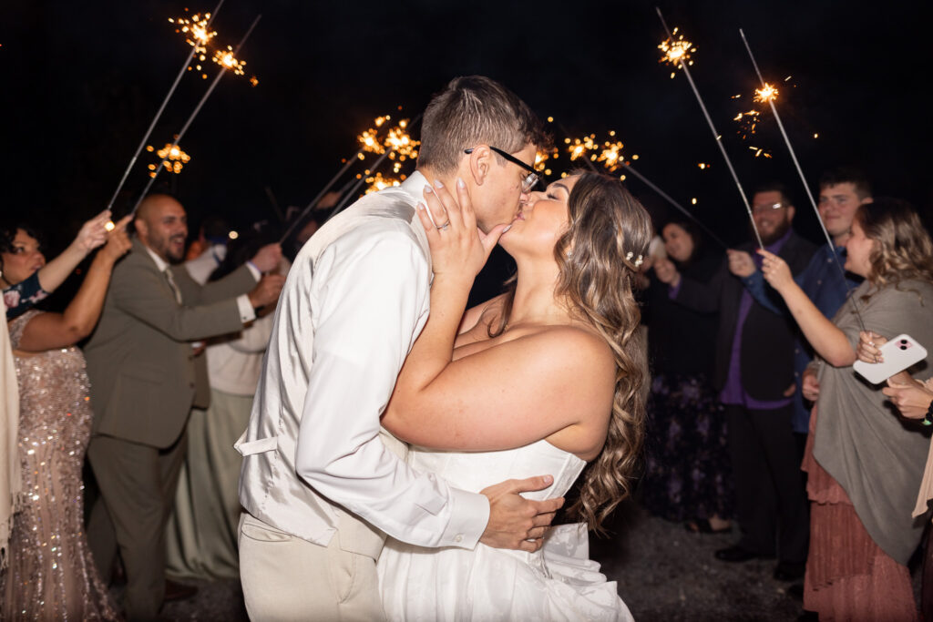 In a close-up view framed by raised sparklers, the groom cups the bride’s face as they kiss deeply, bright golden sparks lighting their faces while guests cheer on both sides during their wedding send off.