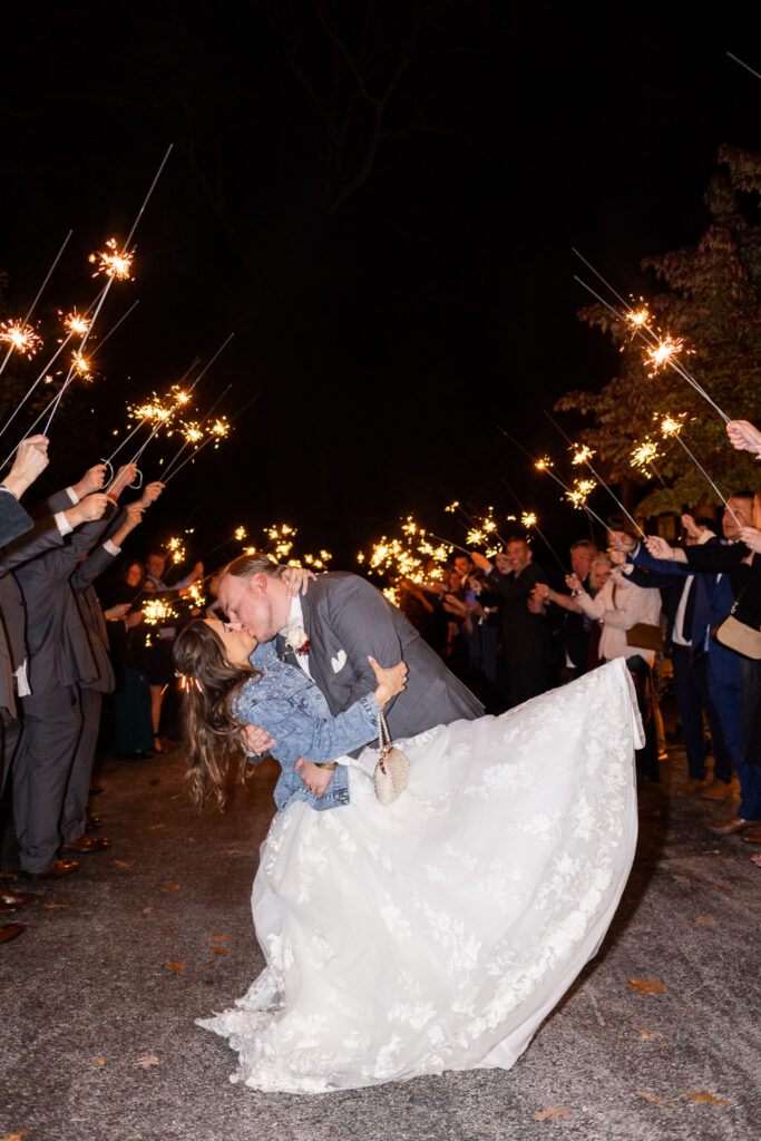 At night on a gravel drive, friends and family hold long sparklers high overhead creating a glowing tunnel as the bride dips backward into a kiss with her groom in the center of their sparkling wedding send off.