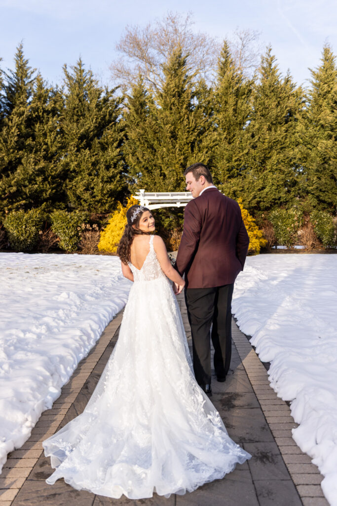 Holding hands, the couple walks away from the camera down a brick path bordered by fresh snow, the bride glancing back over her shoulder while her lace train trails behind, with evergreen trees framing the walkway at a Bucks County wedding venue.