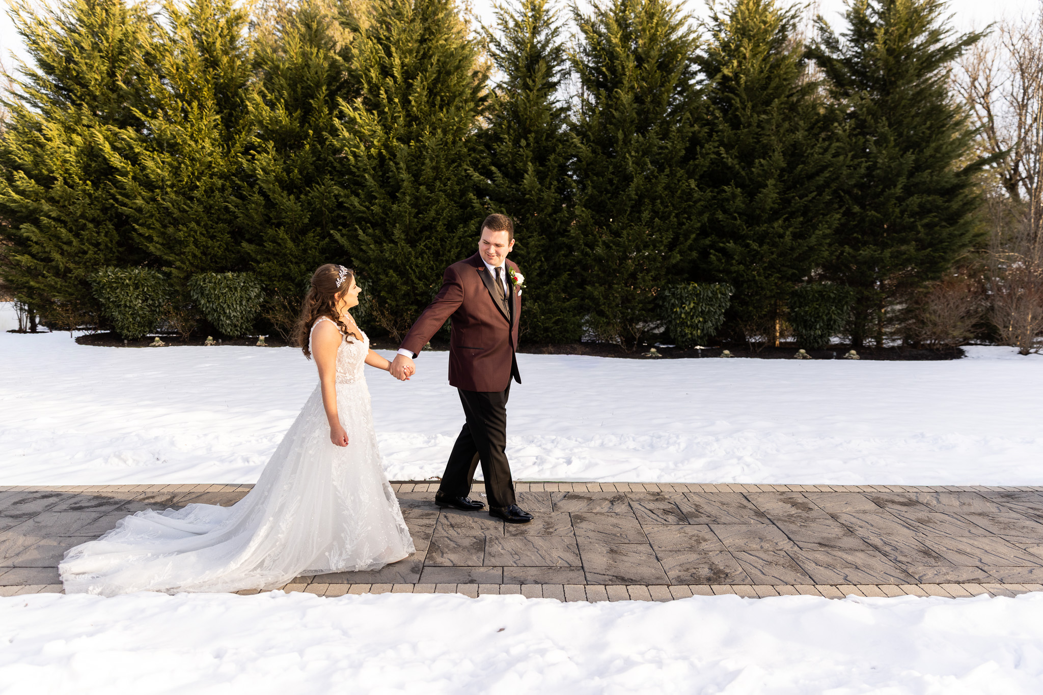 Holding hands, the bride and groom walk together across a paved path bordered by snowbanks and tall evergreen trees, her lace train trailing behind during golden-hour portraits at The Warrington wedding.