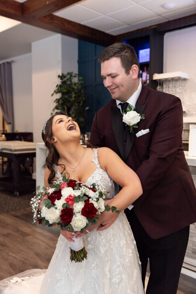 The bride laughs openly as she leans back into the groom’s arms, holding a bouquet of deep red and white roses accented with eucalyptus, while he smiles down at her from behind in a burgundy tuxedo, capturing a playful and affectionate moment during indoor wedding portraits near the bar area.