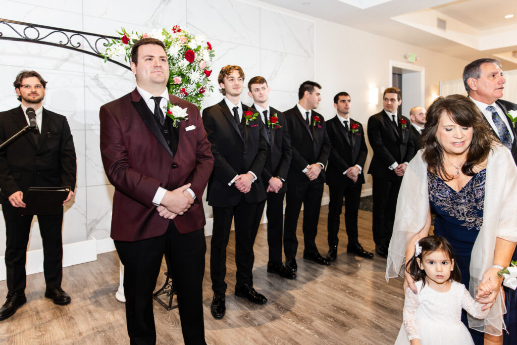 The groom stands at the front of the ceremony in a burgundy suit jacket with black lapels, surrounded by groomsmen in black suits, waiting as the processional begins, conveying anticipation and focus before the bride’s entrance.