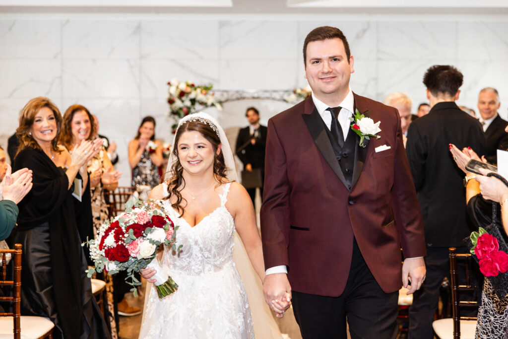 The newly married couple walks back down the aisle hand in hand, smiling broadly as guests stand and applaud around them, capturing the joy of their recessional at a Bucks County wedding venue filled with warmth and celebration.