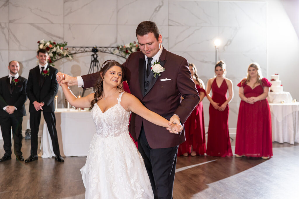 Inside the reception space, the bride and groom share their first dance, arms extended as he guides her into a turn, while their wedding party stands behind them watching, dressed in deep red bridesmaid gowns and black suits during The Warrington wedding.