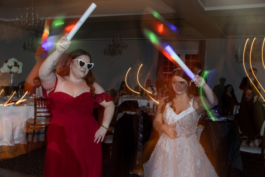 The bride dances beside a bridesmaid wearing heart-shaped sunglasses, both holding glow sticks as colorful light trails streak through the frame, capturing a playful reception moment at a Bucks County wedding venue filled with laughter and movement.