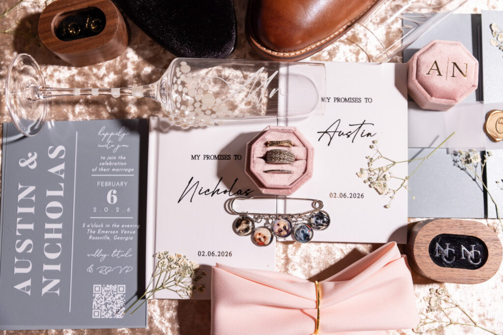 A carefully styled flat lay of LGBT wedding details arranged on a soft champagne velvet backdrop. Two vow books titled “My Promises To Nicholas” and “My Promises To Austin” sit side by side, alongside a grey wedding invitation suite featuring the couple’s names, ceremony date of February 6, 2026, and venue information. A pearl-detailed champagne flute engraved with a flowing “Mr” rests across the stationery. Two pink velvet ring boxes display diamond wedding bands and engagement rings. Brown leather dress shoes, a black velvet loafer, gold cufflinks, a wax seal, a pale blush pocket square secured with a gold ring, and delicate sprigs of baby’s breath complete this elegant same-sex wedding detail composition.