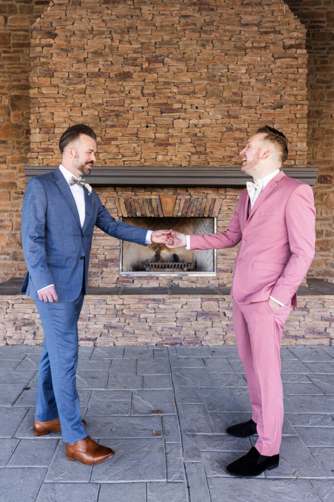 Two grooms stand several feet apart in front of a stacked stone fireplace as they share their first look. Reaching toward one another and smiling mid-laugh. One wears a tailored steel-blue suit with a patterned bow tie and white boutonniere; the other wears a rose-pink suit with a cream bow tie and coordinating vest. Their polished brown and black dress shoes contrast against the gray stone patio floor. The symmetry of the stone hearth behind them creates a timeless portrait that an experienced lgbt wedding photographer captures to highlight connection, movement, and personality during couple portraits.