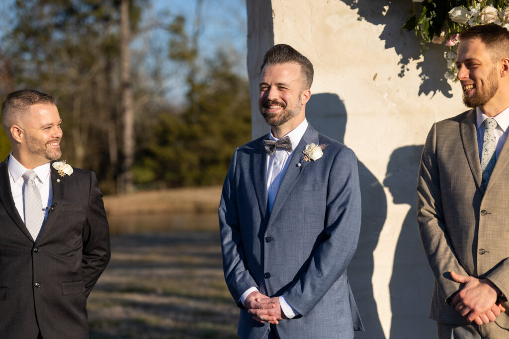 One groom stands at the ceremony site outdoors during golden hour, smiling with visible emotion as he waits. He wears a steel-blue suit with a white boutonniere and patterned bow tie. Two groomsmen in dark suits stand nearby. Soft sunlight casts warm shadows against a white column, framing this heartfelt moment before the LGBT wedding ceremony begins.