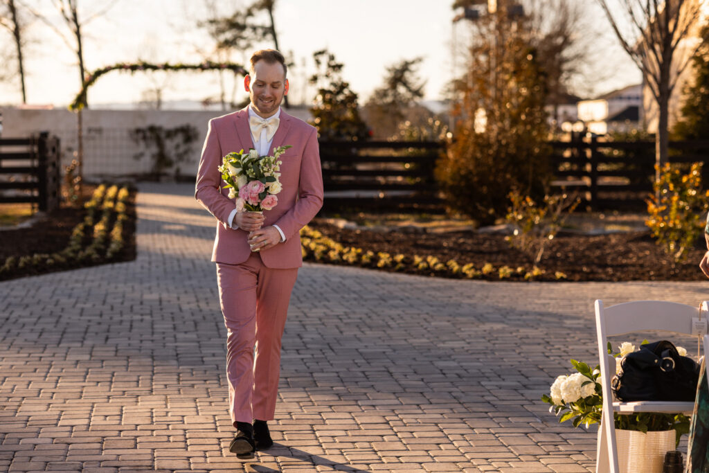 A groom in a tailored pink suit walks along a stone pathway holding a bouquet of blush roses and greenery at a wedding venue in North Georgia. Golden sunlight casts long shadows across the landscaped garden beds and ceremony arch in the background, capturing a peaceful pre-ceremony moment.