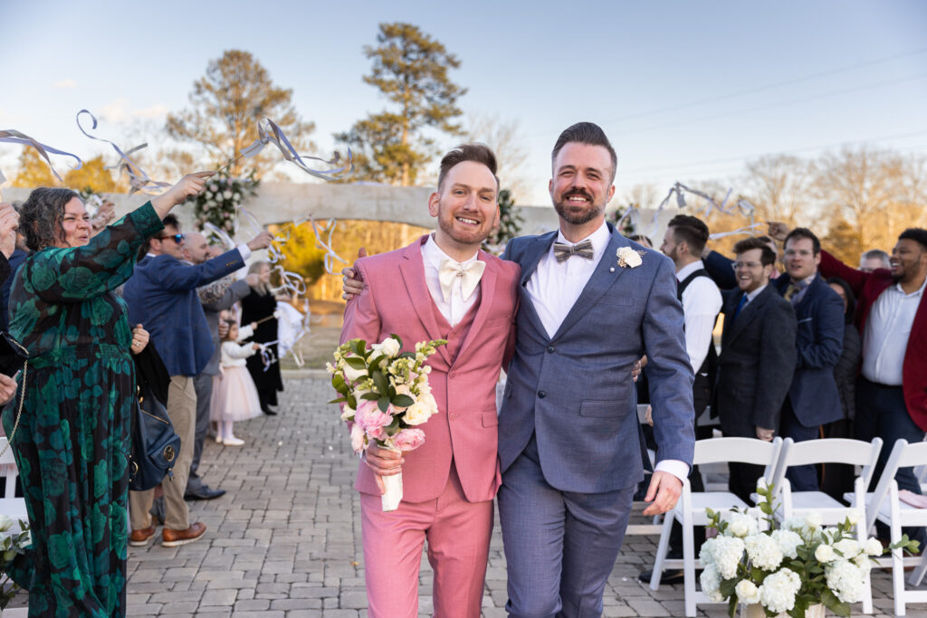 Arm in arm, the grooms walk back down the aisle smiling directly at the camera as guests cheer and streamers flutter around them. The bouquet rests in the pink-suited groom’s hand, and the late-afternoon sunlight casts a golden glow across their faces. This celebratory recessional portrait reflects the candid storytelling style of an lgbt wedding photographer documenting authentic joy.