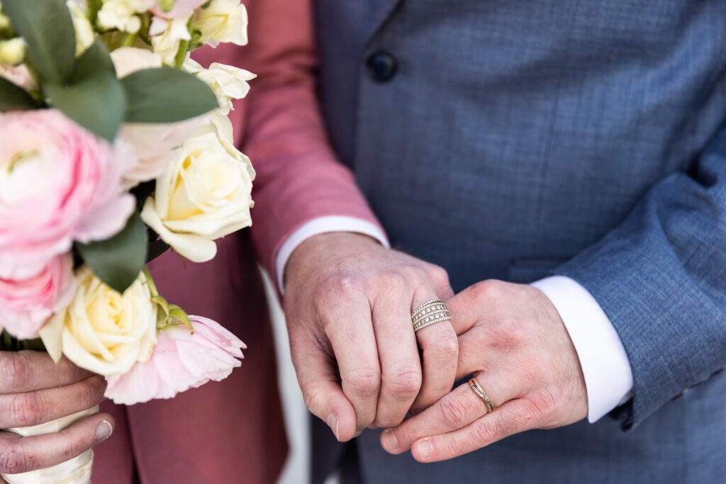 A detailed close-up of the couple’s hands shows matching diamond wedding bands as they gently hold each other. A bouquet of blush and ivory roses with greenery is visible to the side. The pink and blue suit sleeves frame their intertwined fingers, emphasizing commitment and unity in this LGBT wedding portrait.