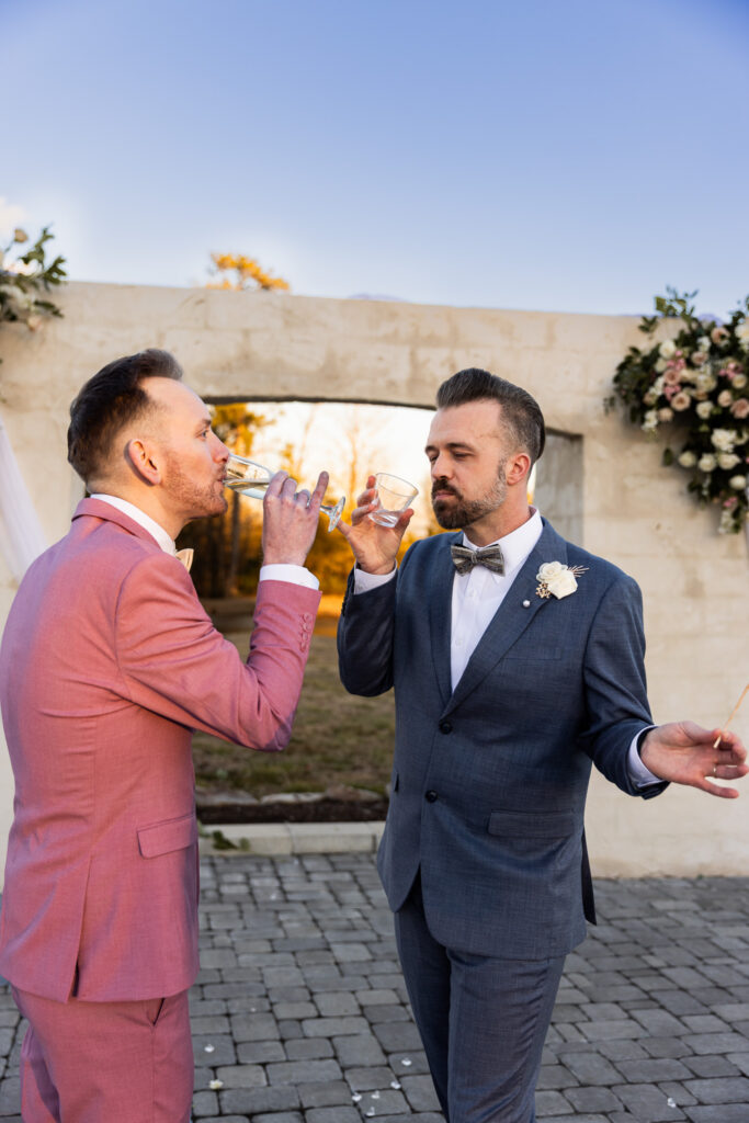 Standing beneath the white ceremony arch at sunset, the grooms raise their glasses mid-toast. The groom in pink sips champagne while the groom in blue lifts a rocks glass, his boutonniere catching the golden light. Floral installations frame them against the soft evening sky. This celebratory moment captures the joy and intimacy that defines heartfelt lgbt wedding photography.