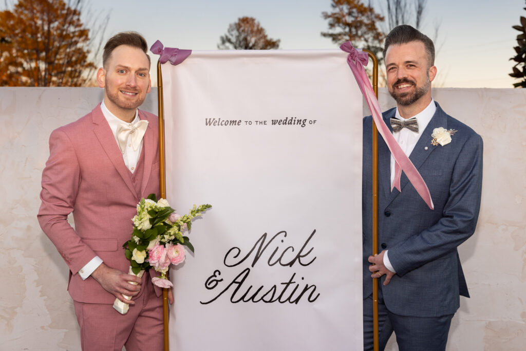 The couple stands beside a framed welcome sign reading “Welcome to the wedding of Nick & Austin,” held between them on a gold stand tied with blush ribbon bows. The groom in pink holds his bouquet, while the groom in blue stands proudly at his side. The clean white backdrop and evening light create a polished portrait that an lgbt wedding photographer captures to document custom wedding signage and personal details.