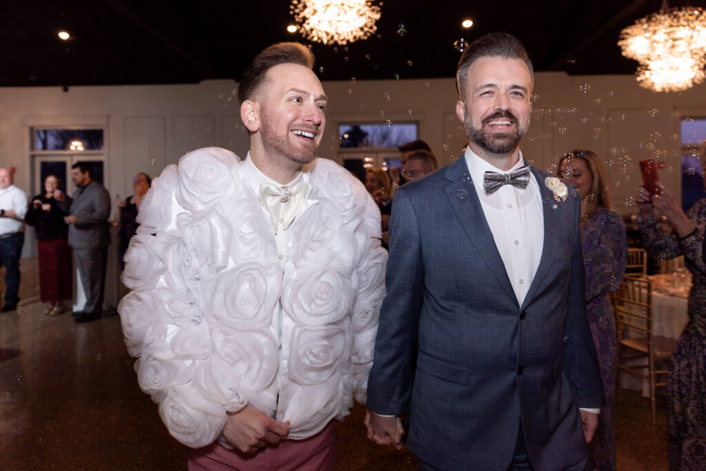 During the wedding reception, the two grooms walk hand in hand through a room filled with guests and floating soap bubbles. One groom wears a dramatic white floral-textured reception jacket with oversized rosette detailing, while the other remains in his steel-blue suit. Guests smile and celebrate in the background under warm chandeliers, capturing a joyful LGBT wedding reception entrance moment.