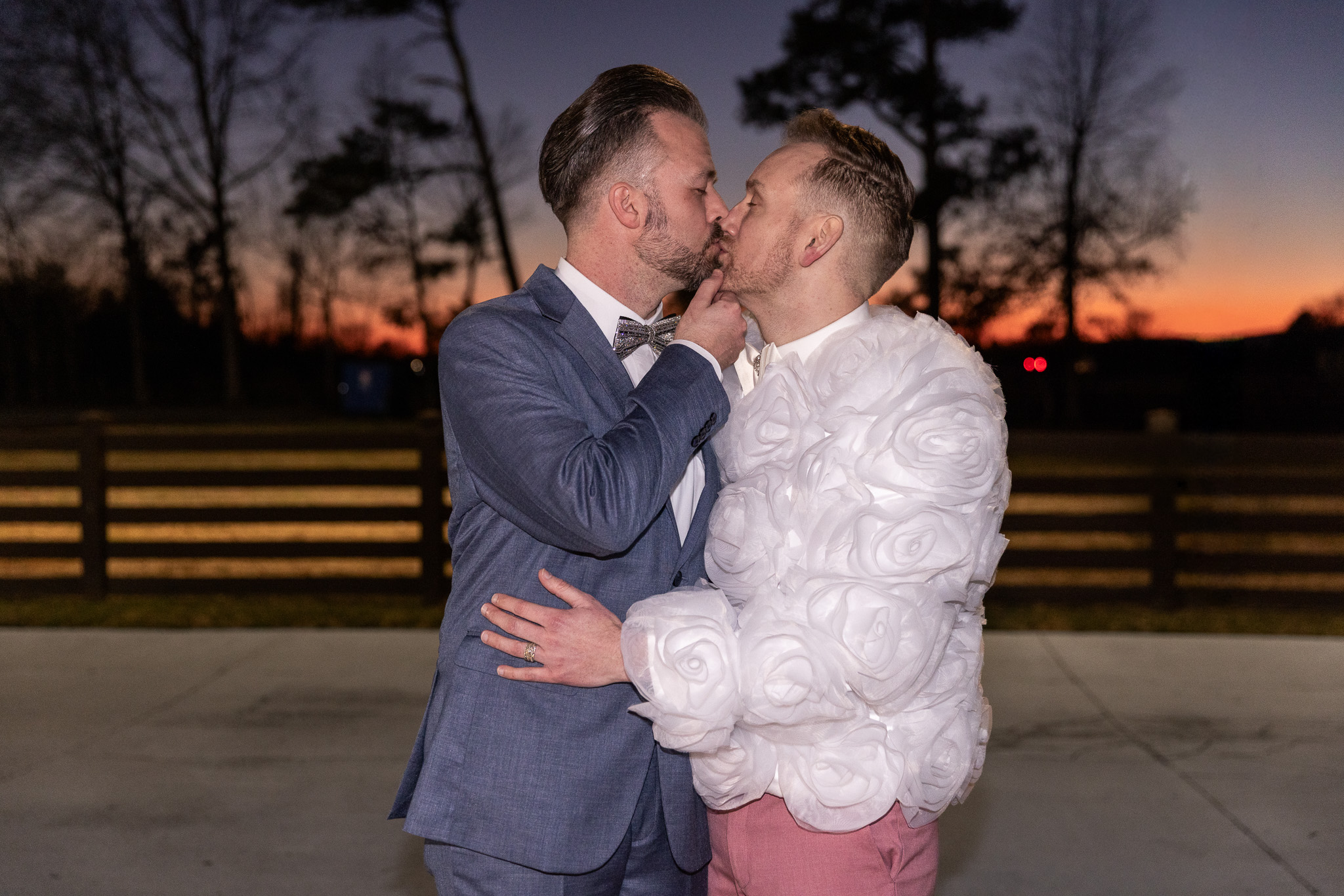 At sunset, the grooms share a kiss outdoors against a dramatic sky glowing with deep orange and purple hues. The groom in pink now wears a bold white reception jacket covered in oversized rosette textures, while the groom in blue remains in his steel-blue suit. A wooden fence and silhouetted trees stretch behind them, creating a cinematic portrait that an lgbt wedding photographer uses to highlight romance during golden hour and twilight.