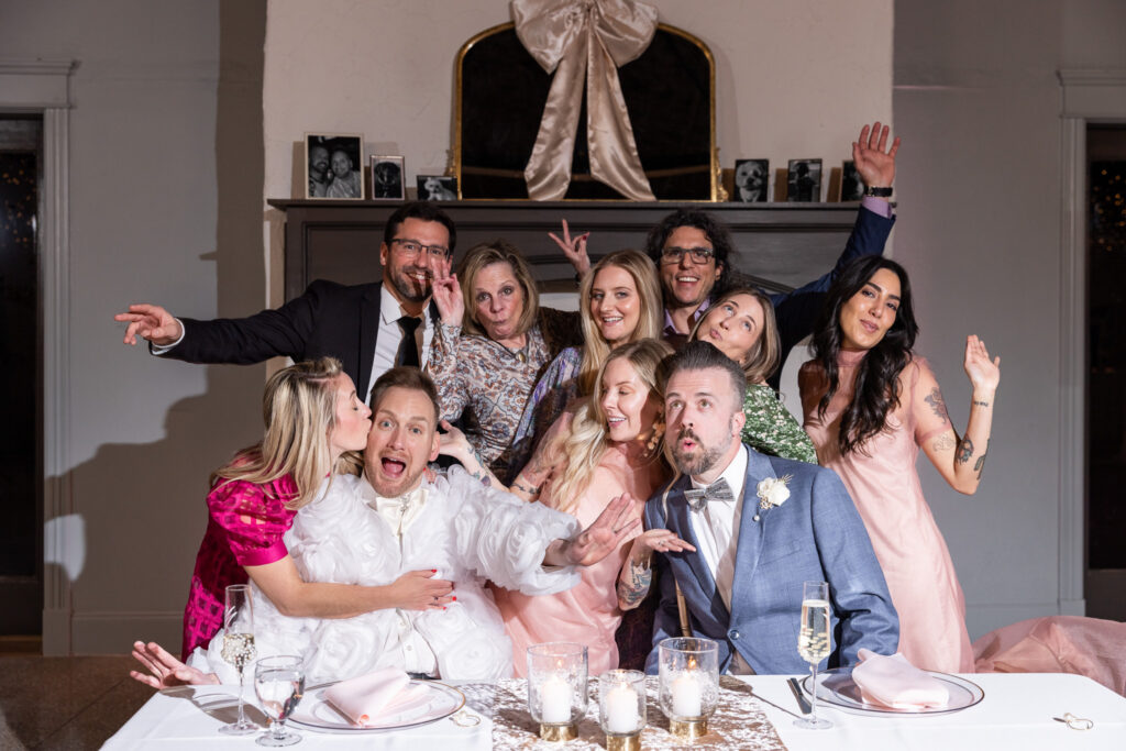 At the sweetheart table, the couple is surrounded by friends striking silly poses for the camera. One friend kisses the groom in pink on the cheek while others throw their hands up in celebration. Champagne flutes, candle votives, and blush napkins decorate the table in front of them. This joyful, personality-filled group portrait embodies the community-centered storytelling that makes lgbt wedding photography so meaningful.