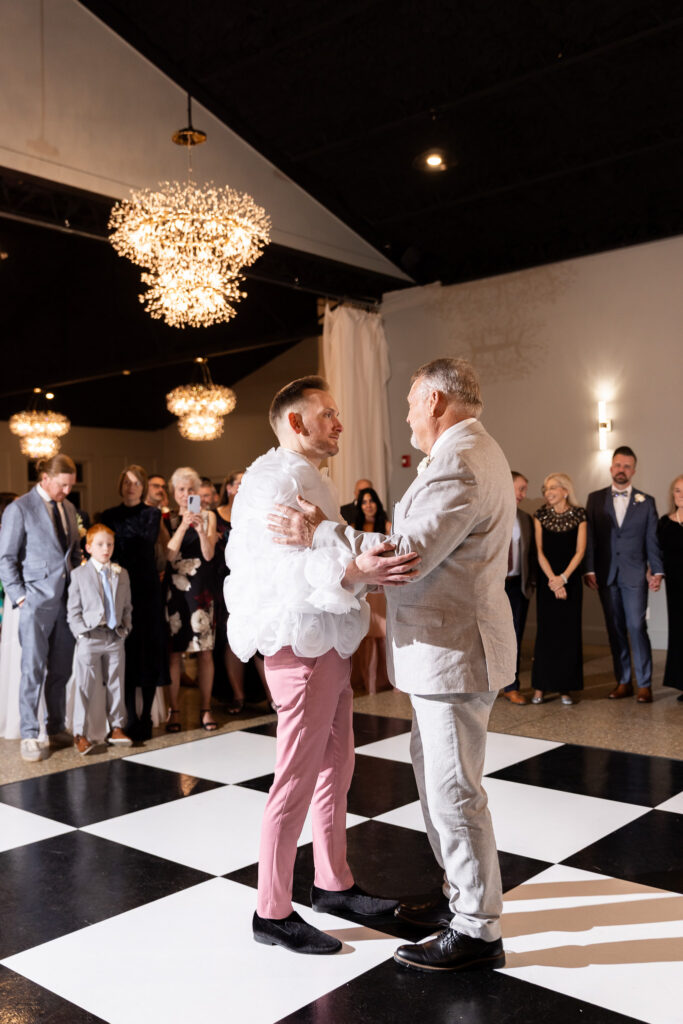 Inside the reception hall beneath sparkling chandelier lights, the groom in pink — now wearing a statement white jacket adorned with oversized rosette textures — shares a slow dance with his father, wearing a light gray suit. Guests stand around the black-and-white checkered dance floor watching tenderly. The emotional family moment is beautifully preserved through documentary-style lgbt wedding photography.